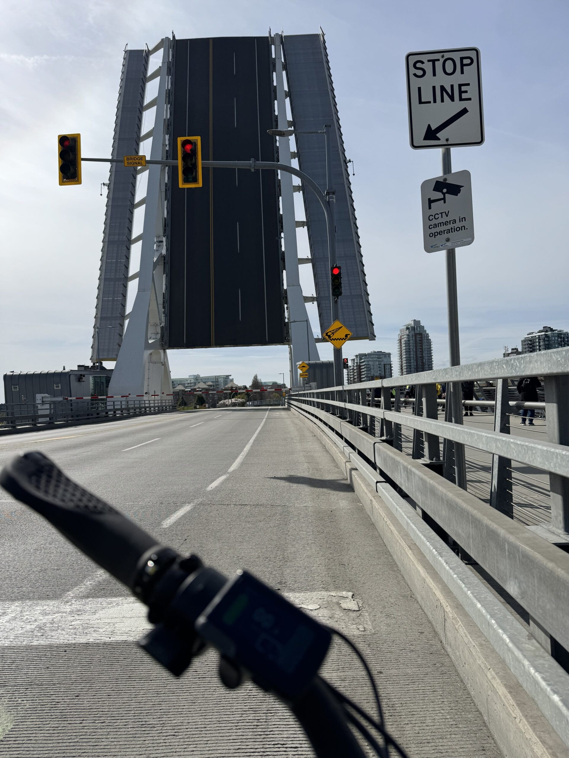 Vue d'un pont basculant levé du point de vue d'un cycliste : guidon de vélo au premier plan, chaussée vide menant à une travée levée verticalement, feux de circulation rouges et panneau “ STOP ” à droite, rambarde métallique pour piétons à droite et bâtiments de la ville au-delà.