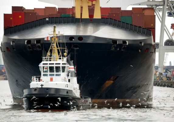 Small tugboat surrounded by flock of seagulls is towing a looming black hulled freighter off the dock