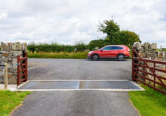 />ALT: Red SUV driving right to left on a paved road at a t-intersection. Viewed through a gate in a fence, with a steel cattle grid.