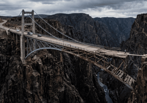 Treacherous canyon with a road leading up to it. Storm clouds are brewing. There is a start of a magnificent bridge that rapidly declines to a rusting steel frame with broken wooden planks, and ends with a single 2x6 wooden plank bridging the final gap. There are stopped cars, one going back, and a man about to walk across the plank.