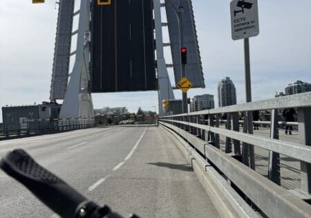 Vista da perspectiva de um ciclista de uma ponte basculante elevada: guidão da bicicleta em primeiro plano, pista vazia que leva a um vão verticalmente levantado, semáforos vermelhos e uma placa de "PARE" à direita, guarda-corpo metálico para pedestres à direita e prédios da cidade ao fundo.